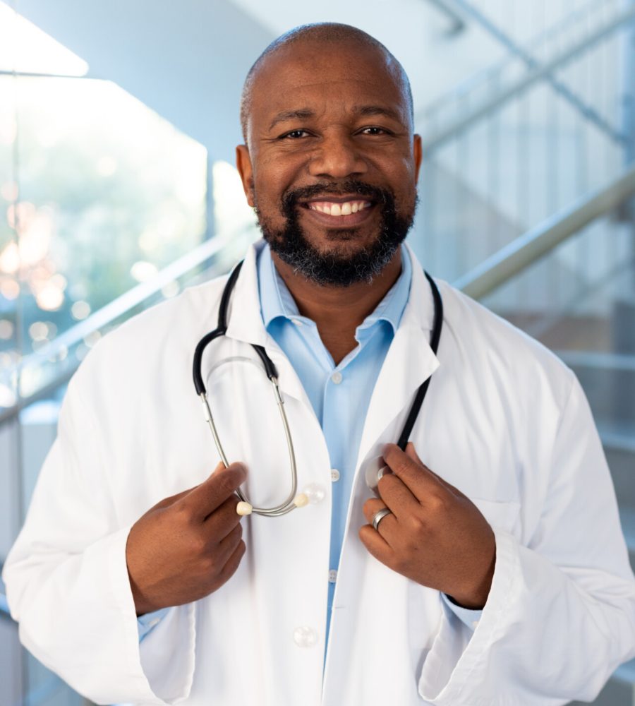 Vertical portrait of smiling african american male doctor in hospital corridor. Hospital, medical and healthcare services.